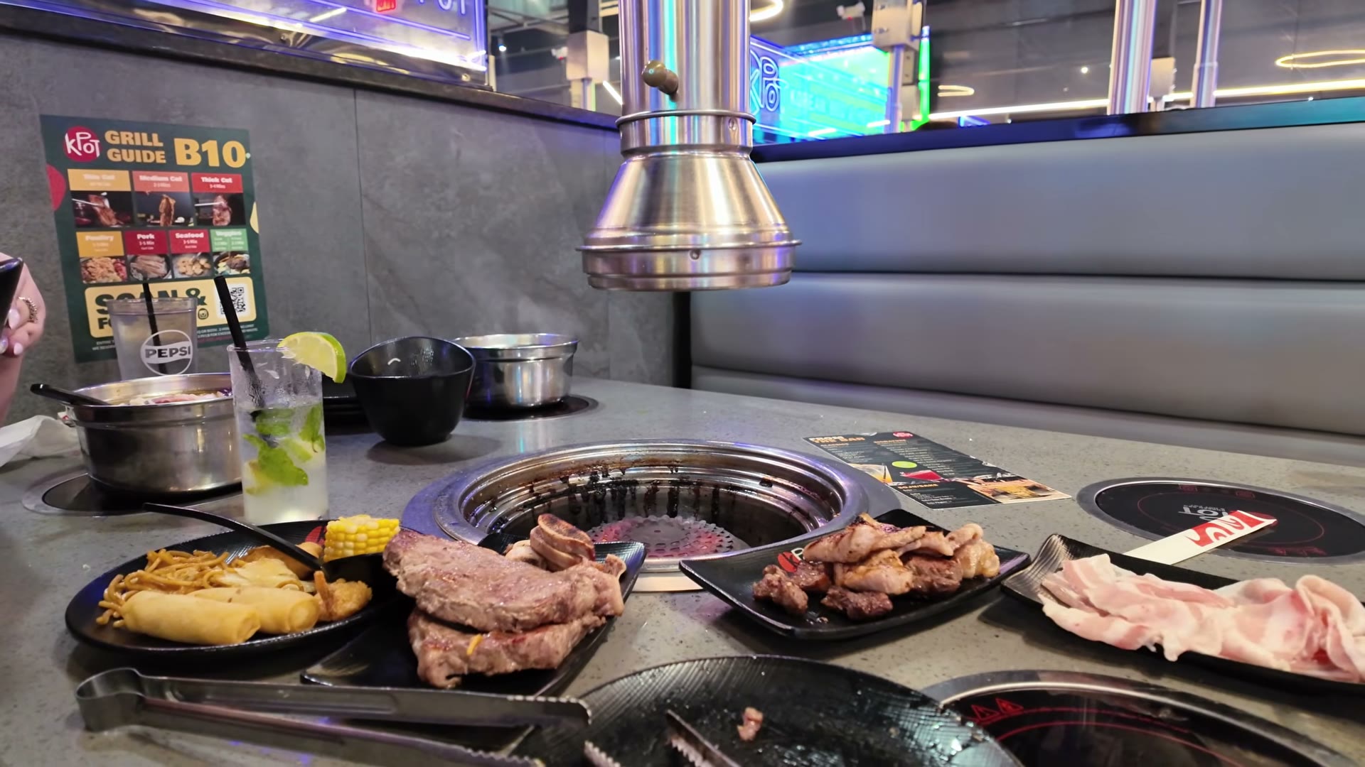 La mesa de KPOT con la parrilla encendida en el centro y las bandejas alrededor: filetes y costillas ya asados, carne a la plancha, panceta cruda, un plato del buffet con fideos, rollitos y mazorca, y un mojito al fondo junto a la olla del hot pot.