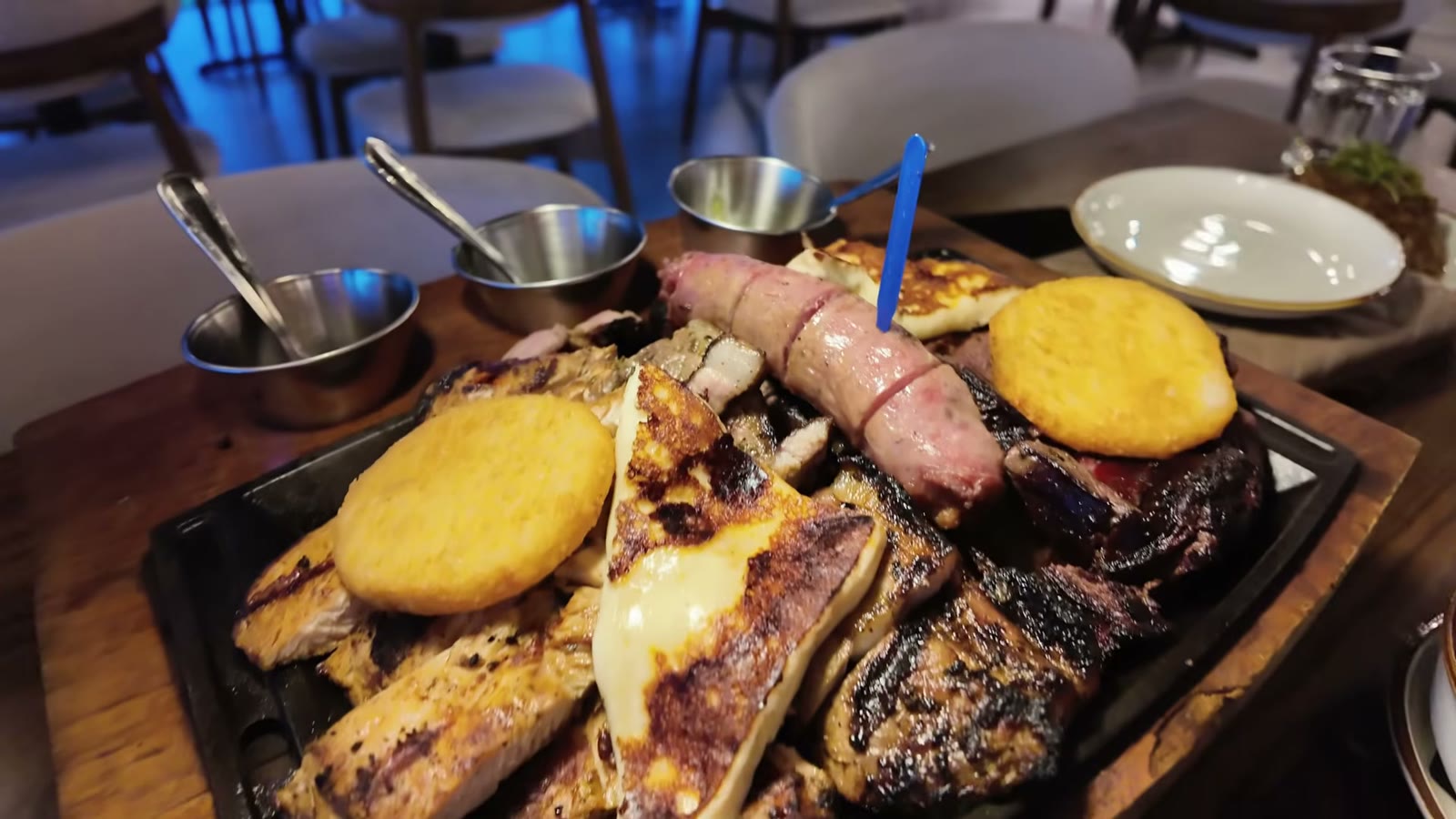La parrilla para compartir de A Fuego Lento sobre su plancha de hierro: cortes a la brasa, chorizo, queso fundido y dos arepitas, con las tres salsas de la casa en cazuelitas al fondo.